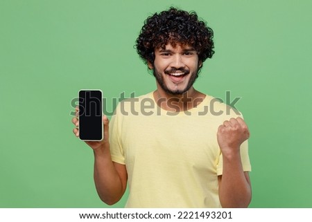 Young excited cool happy Indian man 20s in basic yellow t-shirt hold in hand use mobile cell phone with blank screen workspace area do winner gesture isolated on plain pastel light green background