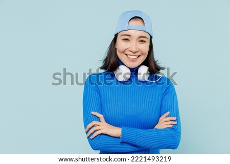 Fancy smiling charming joyful young woman of Asian ethnicity 20s years old wears blue shirt cap headphones around neck hold hands crossed isolated on plain pastel light blue background studio portrait