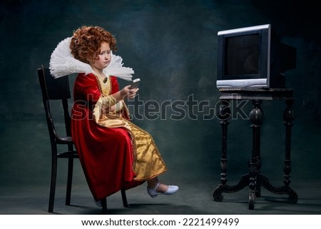 Portrait of little red-headed girl, child, royal person sitting in front of TV isolated over dark green background. Concept of historical remake, comparison of eras, medieval fashion, emotions, queen