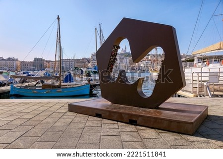 The letter M on the wharf of the Old Port with love locks, boats moored in the marina and the basilica of Notre-Dame de la Garde on the hilltop, Marseille, France