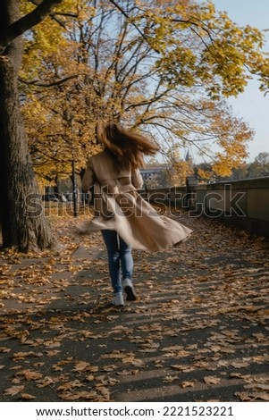 Young woman with long hair in beige trench coat and sneakers running away with her back under the autumn yellow trees on the ground full of fallen leaves in a sunny day. Picture with long exposure.
