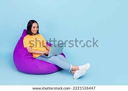 Photo of cheerful adorable nice girl wear yellow t-shirt jeans sit on purple pouf writing laptop isolated on blue color background