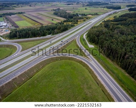 The picture presents intersection of roads visible from above with multiple cars and grass around. 