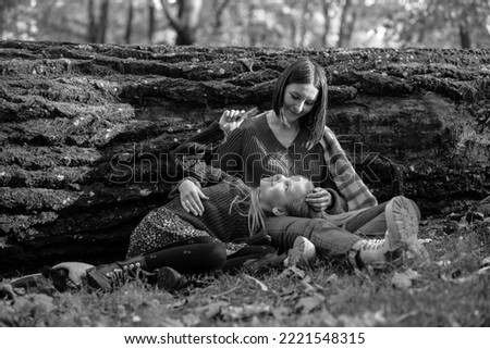 Black and white photo of school aged girl lying on legs of loving mother in warm embraces, felled tree trunk background. Woman and daughter walk in autumn park in cozy clothes. Family, happy moments.