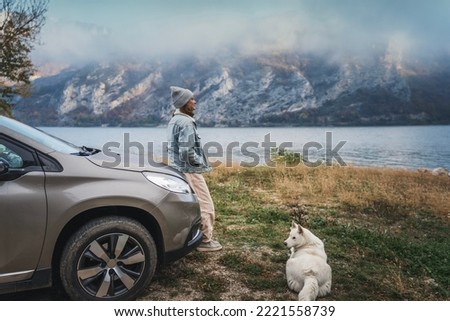 Young woman traveler standing next to the car while traveling with her white swiss shepherd dog on the shore 