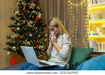 A young woman fell ill during the Christmas holidays. Sitting at home on the sofa near the tree, holding a laptop, consulting with a doctor via video call. He wipes his nose with a napkin, feels bad. Royalty-Free Stock Photo #2221562943