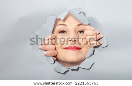 Surprised woman looking playfully in torn paper hole, has excited cheerful expression, looks through breakthrough of gray background. Wow, great news. Closeup