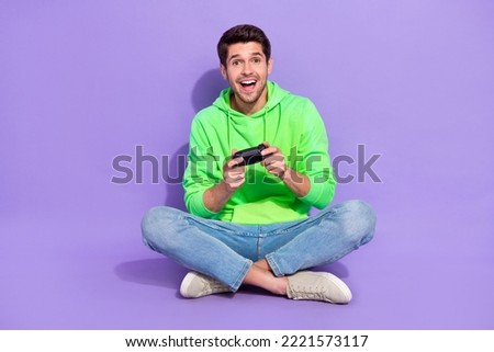 Full length photo of positive excited man wear green hoodie having fun playstaton isolated purple color background