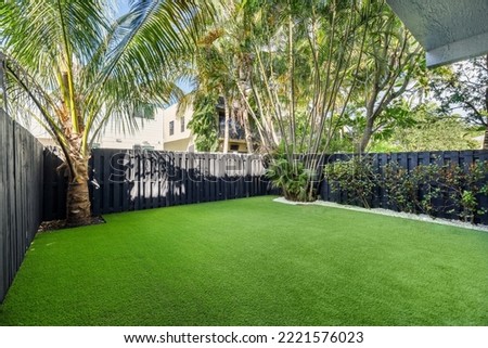Patio with artificial grass, fenced with wooden fences, flowerbed with white stones, tropical plants, palms, buildings in the background