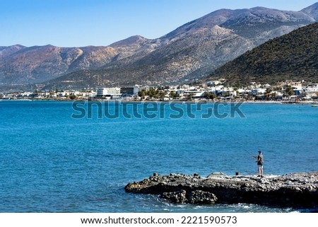 Awesome Picture of a man fishing on the cliff edge. In the background is the beautiful blue Crete sea and coast with mountains and sky.
