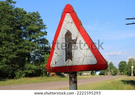 A dirty old rusty triangular road sign with a red frame against a background of green trees and blue sky