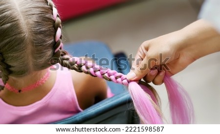 Beautiful blond girl, of seven years old, braided two pigtails, do a hairstyle with pink locks of hair in a beauty salon, a hairdresser's salon, in front of a large mirror. a little princess. High Royalty-Free Stock Photo #2221598587