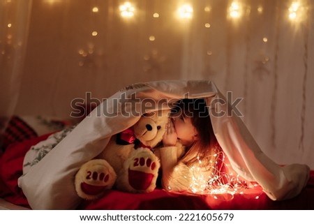 little cute smiling baby girl with white teddy bear peeking out from under a blanket on a bed at home with lighting garlands at dark. Child looking in camera. Fairytale childhood with miracle concept. Royalty-Free Stock Photo #2221605679