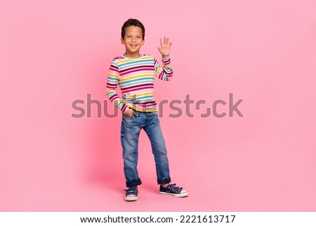Full length photo of positive sweet optimistic boy dressed striped shirt jeans arm in pocket wave palm isolated on pink color background