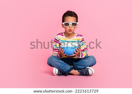 Full size photo of cute impressed boy dressed striped long sleeve jeans sitting hold pop corn bowl isolated on pink color background