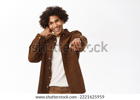 Call me. Happy young guy showing mobile phone gesture and pointing at you, asking your number, standing over white background