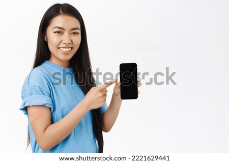 Smiling asian girl with band aid after coronavirus vaccine, shows smartphone screen, covid health certificate on mobile phone, white background