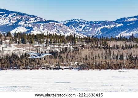 Small bush plane aircraft takes off from frozen Lake Laberge in Yukon Territory winter wilderness landscape of boreal forest taiga hills, YT,  Canada Royalty-Free Stock Photo #2221636415