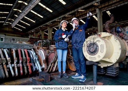 Wide shot of Caucasian factory man use walkie talkie to contact his team and stand with Asian co-worker woman near spare part of machine of the train in industrial business workplace.