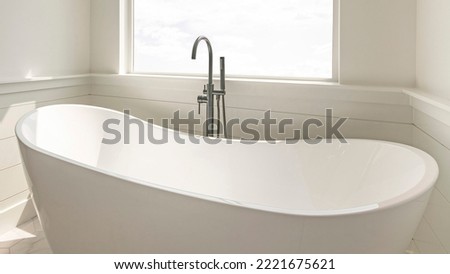 Panorama Modern solid base freestanding tub with floor mount dual faucets against the square picture window. Bathroom interior with marble tiles and shower stall with glass and black fixtures. Royalty-Free Stock Photo #2221675621