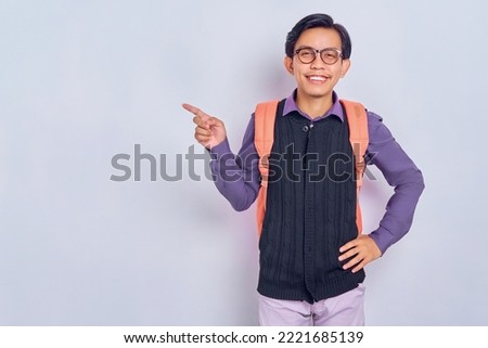 Look here! Smiling handsome young Asian student man with backpack pointing left, space for text, advertising isolated on gray background. Education in high school university college concept