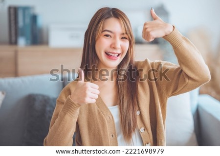 Satisfied asian woman smiling while looking at camera with thumbs up sign sitting on a couch in the living room at home. Body language. I like that Royalty-Free Stock Photo #2221698979