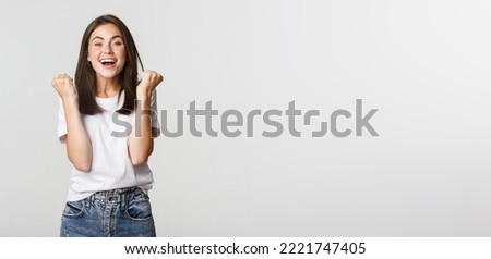 Cheerful attractive brunette girl fist pump and smiling with rejoice, winning. Royalty-Free Stock Photo #2221747405