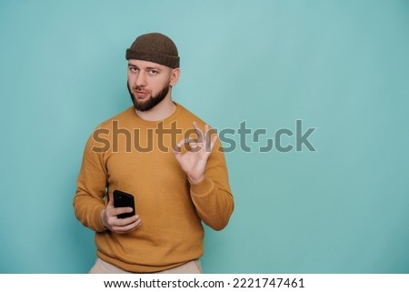 Confident young beardy man in hat and sweater shows sign ok by hand holding phone against turquoise wall at studio looking at camera. Isolated male approved quality. Promo, mockup. Perfect choice.