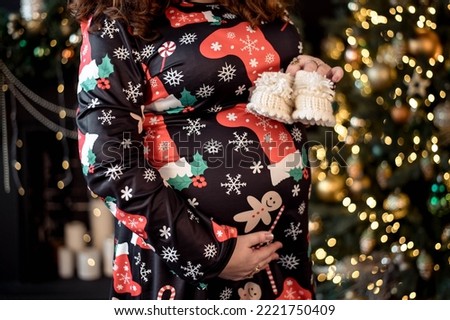 a pregnant woman poses against the background of a Christmas tree, holding booties in her hands