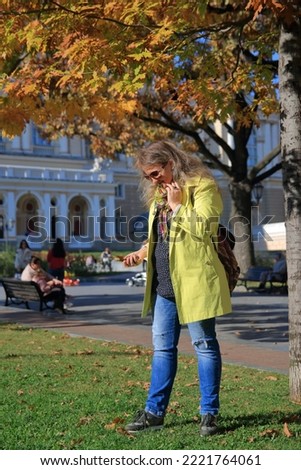 The photo was taken in the city of Odessa in Ukraine. The picture shows a young woman in a yellow raincoat looking for acorns in the green grass of an autumn park.