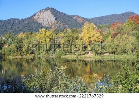 A picture of a stunning autumnal landascape with lots of colors, trees and a romantic lake surrounded by high mountains, location Bulgaria