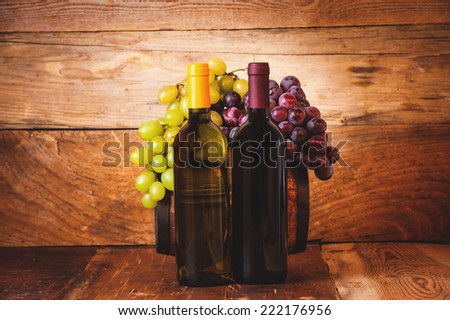 Red and white wine bottle with grapes and barrel on wooden rustic table.