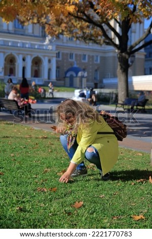 The photo was taken in the city of Odessa in Ukraine. In the picture, a young woman tourist collects oak acorns on green grass in a public autumn park.