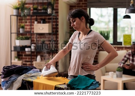 Bored woman tired of ironing clothes at home watching in frustration while doing household chores. African American husband in background resting while wife does all the work. Royalty-Free Stock Photo #2221773223