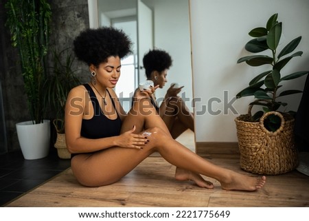 African american woman applying cosmetic cream on knee near mirror and plants in bathroom