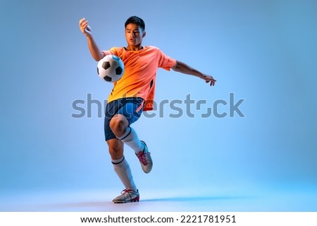 Portrait of young man, football player in motion, action, kicking ball with knee isolated over blue background in neon light. Concept of sport, team game, action, motion. Copy space for ad, poster Royalty-Free Stock Photo #2221781951