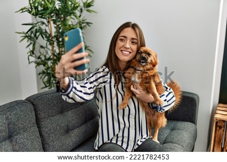 Young hispanic woman make selfie by the smartphone sitting on sofa hugging dog at home