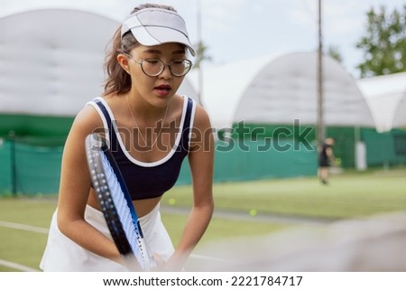 Beautiful girl of Asian appearance is preparing for tennis match. Student in glasses holds racket in hands, wears tennis cap, closes eyes and prepares to make serve. She is dressed in top and skirt. Royalty-Free Stock Photo #2221784717