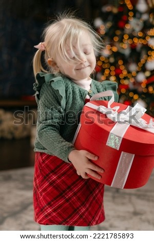 Little blonde girl holds big gift. Portrait of fair-haired child with red gift box. Vertical frame. New Year mood