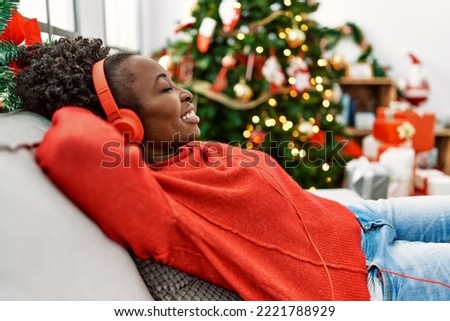 African american woman listening to music sitting on sofa by christmas tree at home Royalty-Free Stock Photo #2221788929