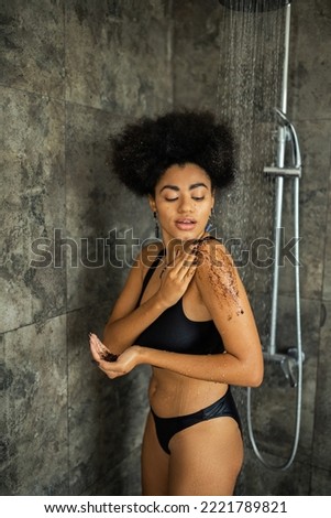 Pretty african american woman applying coffee scrub on shoulder in shower