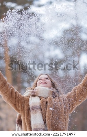 A white blonde girl in a knitted hat, a long scarf and a fur coat walks through the forest and smiles. Woman catches snow in white mittens. Winter is on the street. High quality photo.