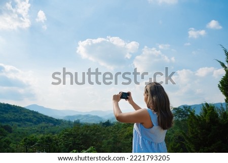 A beautiful young Asian woman wearing a blue dress uses a smartphone to take pictures of the mountain landscape, evening sky, clouds on a mountain full of trees. Lifestyle, leisure and travel