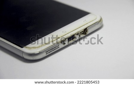 Old and used smart phone mobile device. Technology cellphone on white plain background. Diagonal view, bottom part of the device with damaged screen protector glass and charger port and earphone hole