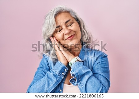 Middle age woman with grey hair standing over pink background sleeping tired dreaming and posing with hands together while smiling with closed eyes. 