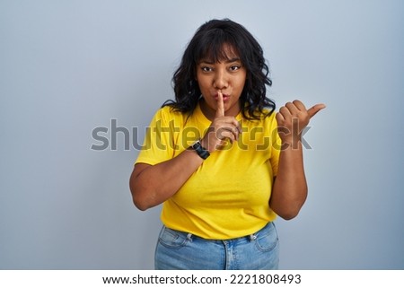 Hispanic woman standing over blue background asking to be quiet with finger on lips pointing with hand to the side. silence and secret concept. 