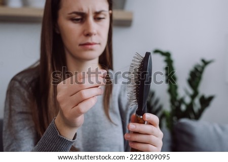 Selective focus of young girl sitting on sofa brushing hair with hair tool, worrying about hair loss shedding or bad condition. Upset brunette woman styling hair with comb, hormonal imbalance concept