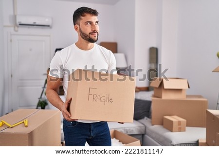 Handsome hispanic man moving to a new home holding fragile box smiling looking to the side and staring away thinking. 