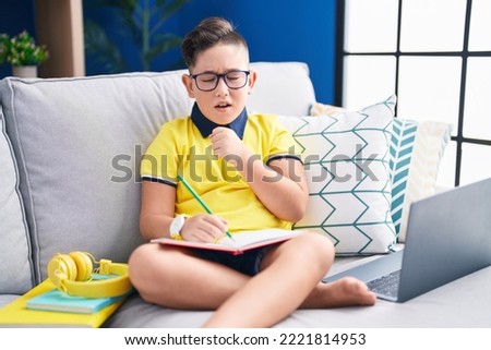 Young hispanic kid doing homework sitting on the sofa feeling unwell and coughing as symptom for cold or bronchitis. health care concept.  Royalty-Free Stock Photo #2221814953