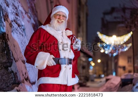 Santa Claus on Xmas Eve at city streets. Friendly cheerful outgoing senior man with real white beard Father Christmas at snowy old town lifestyle scenery. New Year, celebration, garlands illumination